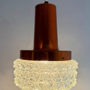 May include: A retro pendant light with a copper-coloured metal top and a clear, textured glass shade. The shade features a geometric pattern, and the light is suspended by a white cord. This vintage lighting fixture is a stylish addition to any room.