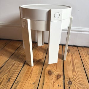 May include: A cream-coloured side table with a geometric design. The table has a circular top, a cylindrical mid-section, and four angled legs. The table is made of a solid, opaque material and stands on a wooden floor.