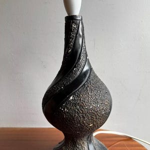May include: A black, textured table lamp with a white bulb socket. The lamp has a curvy design with a textured surface and a glossy black stripe. The lamp is on a wooden surface.