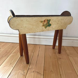 May include: A vintage wooden planter with a cream-coloured body and a painted rose design. The planter has four brown legs and curved handles on each end. The planter is approximately 61 cm long.