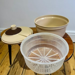 May include: A retro salad spinner set, featuring a cream and brown lid, a beige bowl, and a white plastic basket. The set is displayed on a small wooden table with black legs, evoking a vintage kitchenware style.