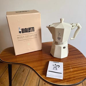 May include: A cream-coloured Bialetti Moka Exclusive stovetop espresso maker with an octagonal base, a matching cream box labelled Moka Exclusive, and a small instruction booklet featuring a graphic of the coffee maker on the front cover.