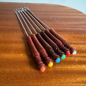 May include: A set of six fondue forks. Each fork has stainless steel tines and a wooden handle. The handles have coloured tips: red, orange, yellow, green, blue, and white. The forks are on a wooden surface.