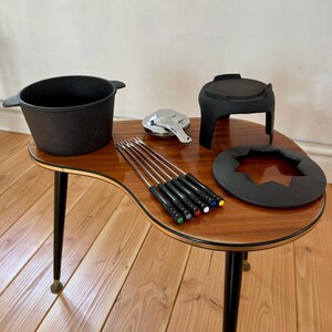 May include: A vintage fondue set comprising a black pot, stand, and burner. The set includes six fondue forks with coloured tips and a metal lid. The items are arranged on a wooden table with black legs.