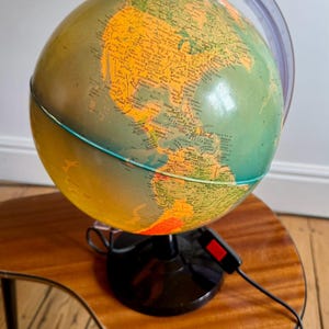 May include: Illuminated globe in green and yellow, displaying geographical features. The globe is on a black base with a red power switch. It rests on a wooden table with a distinctive, curved shape, adding a vintage touch.