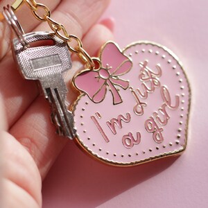 May include: A pink heart-shaped keychain with a gold border and a pink bow. The keychain has the text "I'm just a girl" written on it.