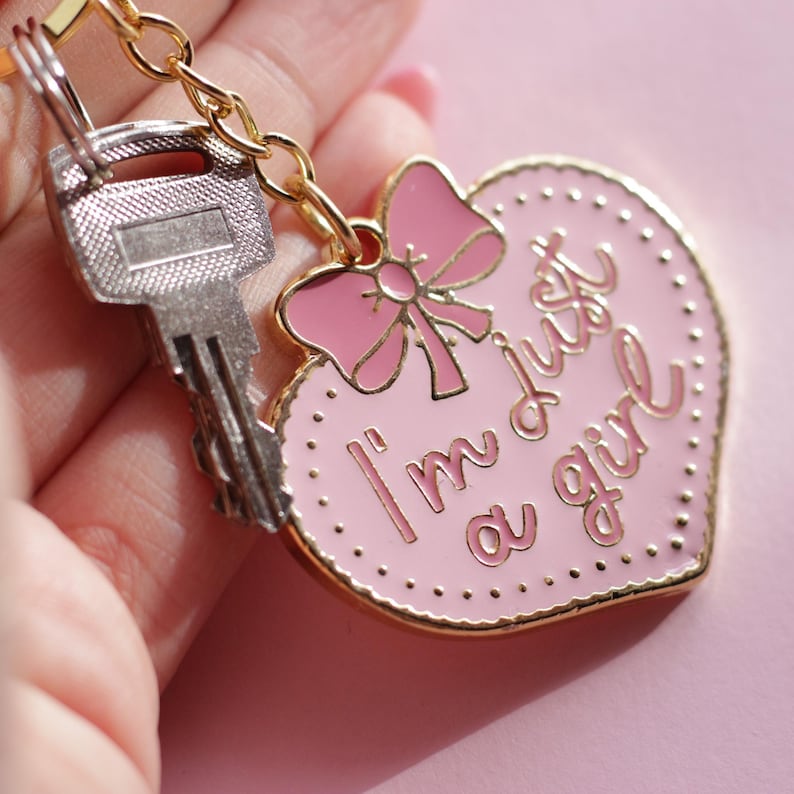May include: A pink heart-shaped keychain with a gold border and a pink bow. The keychain has the text "I'm just a girl" written on it.