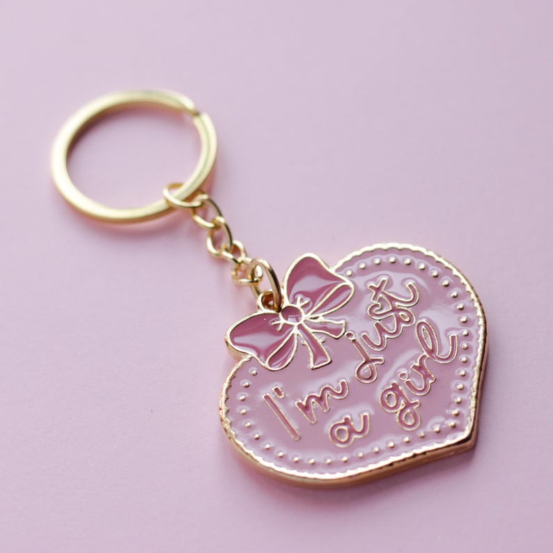 May include: Pink heart-shaped keychain with a gold chain and a pink bow. The heart has a gold border and the text "I'm just a girl" in gold lettering.