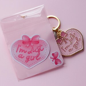 May include: A pink heart-shaped keychain with a gold metal frame and the text "I'm just a girl" in pink lettering. The keychain comes with a sticker of a pink heart with a bow and the same text.