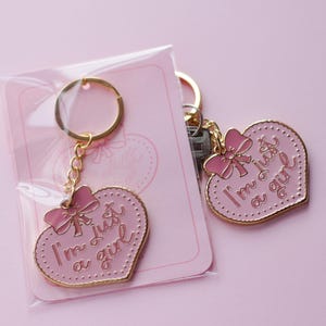 May include: Two gold heart-shaped keychains with pink enamel and gold accents. The keychains feature a pink bow and the text "I'm just a girl".