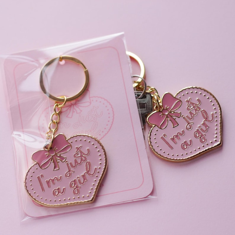 May include: Two gold heart-shaped keychains with pink enamel and gold accents. The keychains feature a pink bow and the text "I'm just a girl".