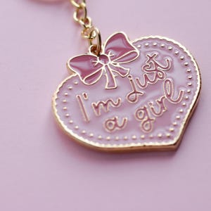May include: Pink heart-shaped keychain with a gold border and a pink bow. The keychain has the text "I'm just a girl" written on it.