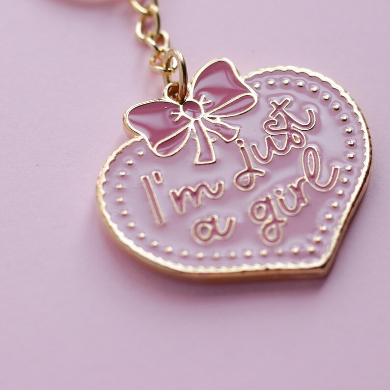 May include: Pink heart-shaped keychain with a gold border and a pink bow. The keychain has the text "I'm just a girl" written on it.