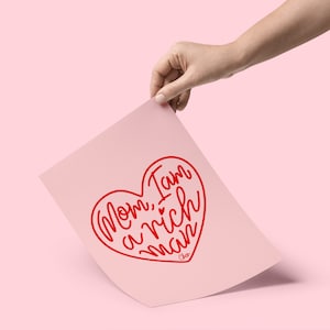 Pode incluir: Um cartão rosa com um coração vermelho contendo o texto "Mãe, eu sou um homem rico". O cartão está sendo segurado por uma mão.