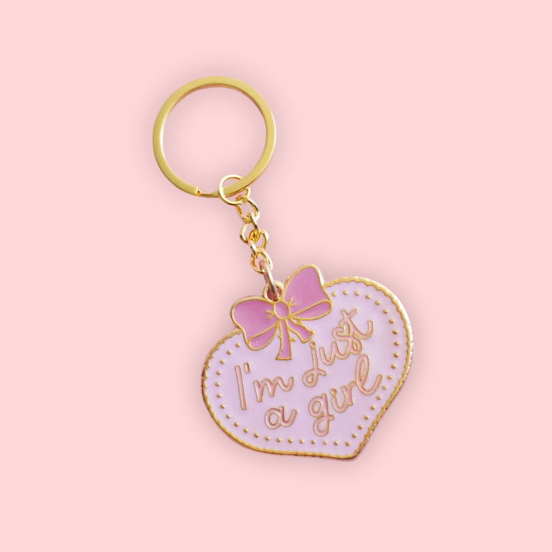 May include: A pink heart-shaped keychain with a gold chain and a pink bow. The keychain has the text "I'm just a girl" written on it.
