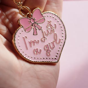 May include: Pink heart-shaped keychain with gold trim and a pink bow. The keychain says "I'm just a girl" in gold lettering.