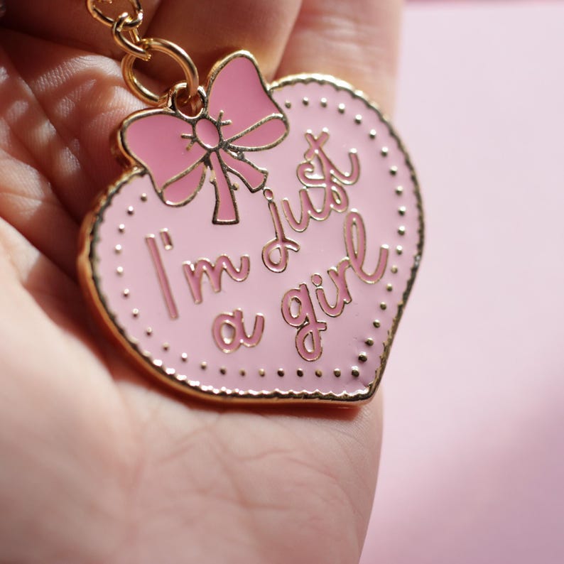 May include: Pink heart-shaped keychain with gold trim and a pink bow. The keychain says "I'm just a girl" in gold lettering.