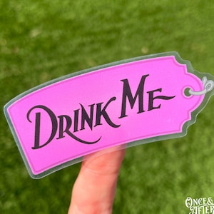 May include: A pink tag-shaped sticker with the words "DRINK ME" in black script. The sticker has a clear border and a small hole at the top. The sticker is held up by a finger against a green background.
