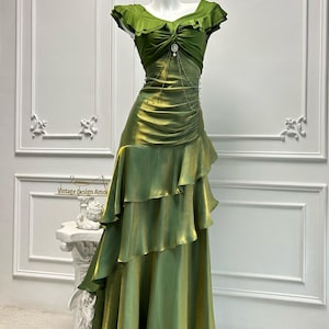 May include: A green satin gown on a mannequin. The dress features a ruffled neckline and a tiered skirt. A decorative embellishment is at the waist. The dress is displayed in front of a white wall. The text "Vintage Design Amo" is visible.