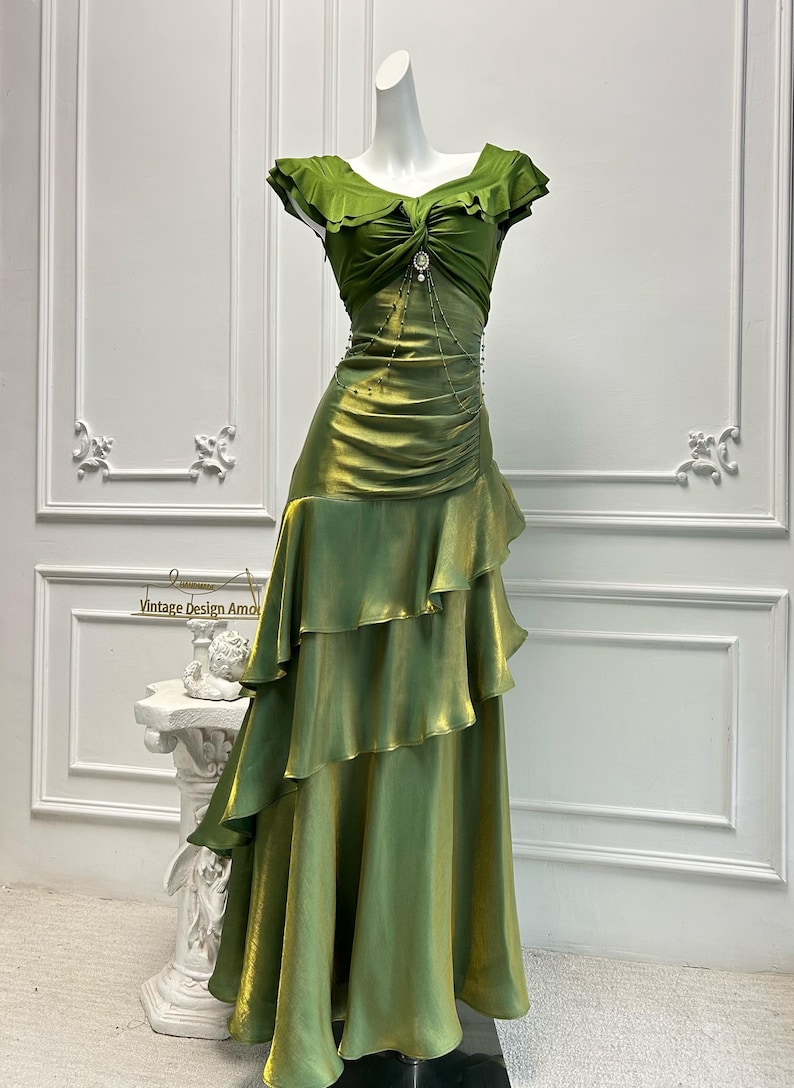 May include: A green satin gown on a mannequin. The dress features a ruffled neckline and a tiered skirt. A decorative embellishment is at the waist. The dress is displayed in front of a white wall. The text "Vintage Design Amo" is visible.