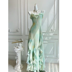 May include: A flowing, floor-length gown in shades of mint green and pale yellow, displayed on a mannequin. The dress features delicate ruffled accents and a floral detail at the neckline. A necklace adorns the mannequin's neck. The dress is a formal wear item.