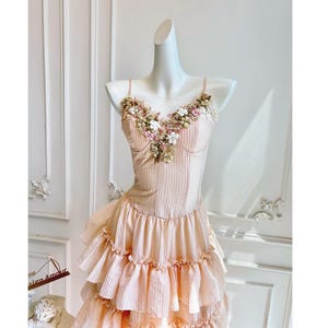 Peut inclure: Robe pêche claire sur mannequin. La robe présente un corsage ajusté orné de fleurs et une jupe à volants superposés. Elle est dotée de fines bretelles et d'un motif à rayures verticales.