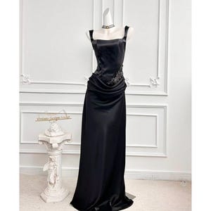Könnte beinhalten: Schwarzes Abendkleid aus Satin auf einer Schaufensterpuppe. Das Kleid hat einen quadratischen Ausschnitt, Spaghettiträger und ein eng anliegendes Oberteil mit verzierten Details. Der Rock fällt bis zum Boden. Ein kleiner dekorativer Ständer mit dem Text "Vintage Design Ambi" befindet sich im Hintergrund.