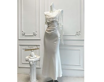 May include: A white satin gown on a mannequin. The dress features a draped bodice with delicate embellishments and a flowing skirt. A sheer, ruffled overlay extends from the shoulder. The background is a white wall with decorative molding.