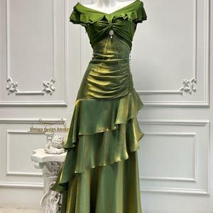 May include: A green, off-the-shoulder formal gown with a layered skirt. The bodice features a ruched design and a decorative embellishment. The dress is displayed on a mannequin against a white background. The text "Vintage Design Amour" is visible.