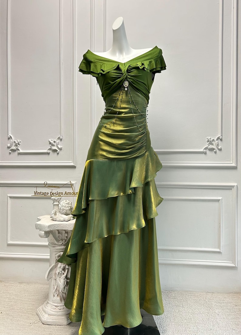 May include: A green, off-the-shoulder formal gown with a layered skirt. The bodice features a ruched design and a decorative embellishment. The dress is displayed on a mannequin against a white background. The text "Vintage Design Amour" is visible.