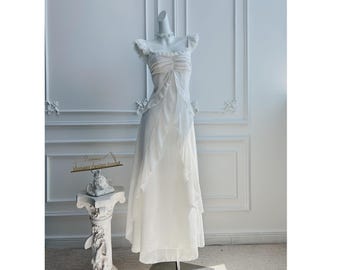 Op de afbeelding: Een lange, witte jurk met gefranjerde kapmouwen en een zwierige rok, gepresenteerd op een mannequin. De jurk staat tegen een witte muur. Een kleine decoratieve standaard met een sculptuur staat op de voorgrond. De jurk heeft een delicate, etherische kwaliteit.