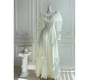 May include: A vintage-style ivory dress on a mannequin. The dress features long sleeves with ruffled cuffs, a high neckline with delicate beading, and a flowing skirt with lace trim. The dress is displayed in a well-lit room with ornate wall paneling.