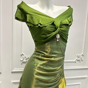 May include: A green off-the-shoulder dress with a ruched bodice and a flowing skirt. The dress features a decorative brooch and a delicate beaded chain. The fabric has a subtle sheen, and the dress is displayed on a mannequin.