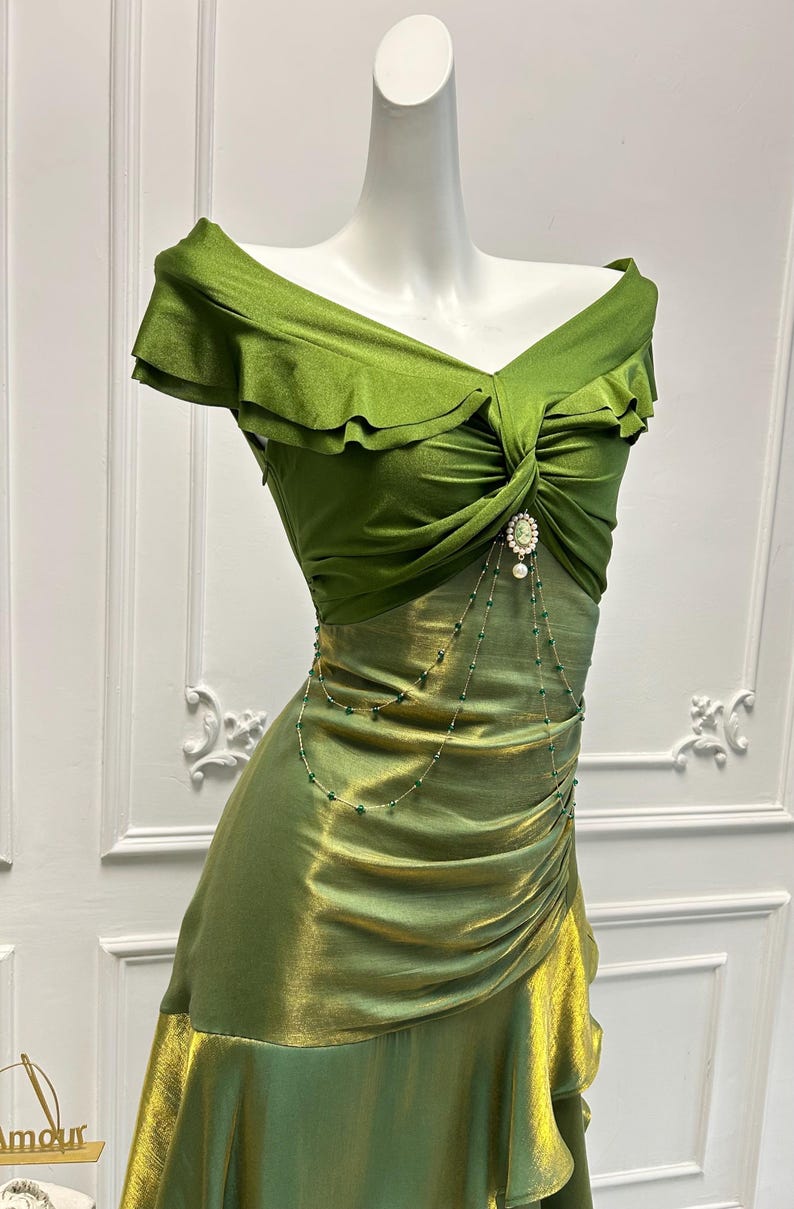 May include: A green off-the-shoulder dress with a ruched bodice and a flowing skirt. The dress features a decorative brooch and a delicate beaded chain. The fabric has a subtle sheen, and the dress is displayed on a mannequin.