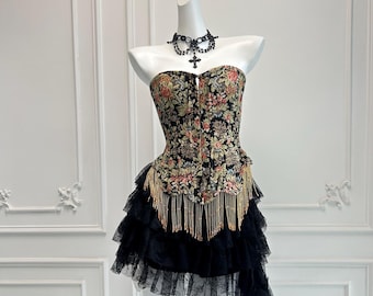 May include: A black strapless corset with a floral tapestry design and gold bead fringe. The corset is paired with a black tulle skirt with multiple layers and a long train.