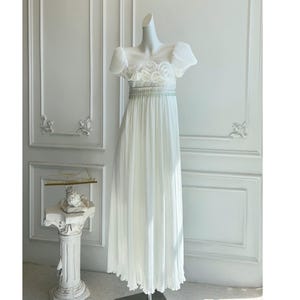 Puede incluir: Un vestido blanco largo hasta el suelo con una falda plisada y mangas cortas abullonadas. El corpiño presenta bordados florales y una cintura con cuentas. El vestido se exhibe en un maniquí contra una pared blanca.