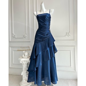 May include: A navy blue formal gown with a sweetheart neckline and spaghetti straps. The bodice is ruched, and the skirt features layered ruffles. A small embellishment is on the side of the bodice. The dress is displayed on a mannequin.