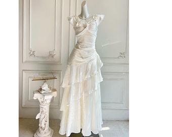 Op de afbeelding: Een witte satijnen jurk met een gelaagde, gerimpelde rok en een gerimpeld lijfje. De jurk heeft kapmouwen en een delicate, met kralen bezette halslijn. De jurk wordt op een mannequin gepresenteerd tegen een witte muur.