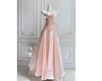 May include: A light pink formal gown on a mannequin. The strapless bodice features floral accents and a flowing skirt. A matching pink necklace is also visible. The dress is displayed in front of a white wall with decorative molding.