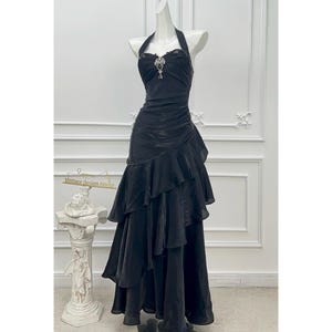 May include: A black floor-length halter dress featuring a sweetheart neckline with a central decorative brooch, a ruched bodice, and a skirt with multiple cascading ruffled layers that create a voluminous, asymmetrical silhouette.