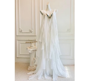 Puede incluir: Un vestido blanco sin mangas hasta el suelo hecho de tela transparente en capas con un escote en V profundo. El escote está adornado con un detalle decorativo con acentos azules, verdes y dorados. La falda consta de varios paneles fluidos.