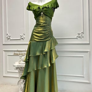 May include: A green off-the-shoulder gown with a layered skirt and a jeweled accent at the waist. The dress has a ruffled neckline and a flowing, tiered skirt. The dress is displayed on a mannequin.