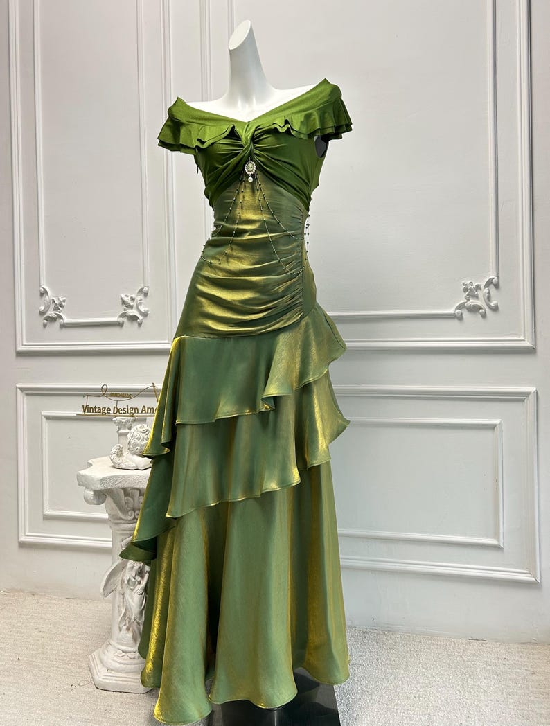 May include: A green off-the-shoulder gown with a layered skirt and a jeweled accent at the waist. The dress has a ruffled neckline and a flowing, tiered skirt. The dress is displayed on a mannequin.
