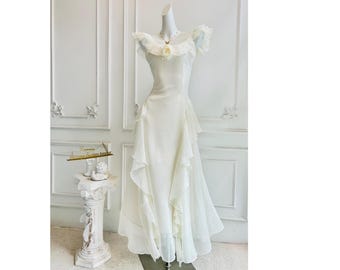 Puede incluir: Vestido de novia blanco de estilo vintage sobre un maniquí. El vestido presenta un escote con volantes, una falda en capas y un delicado adorno floral. El vestido se exhibe frente a una pared blanca con molduras decorativas.