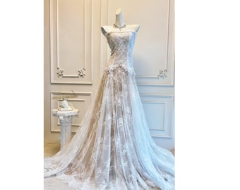 Pode incluir: Vestido de noiva de renda branca sem alças em um manequim. O vestido apresenta detalhes intrincados de renda floral e uma saia fluida. Um colar de prata adorna o pescoço do manequim. O vestido é exibido em uma sala bem iluminada.