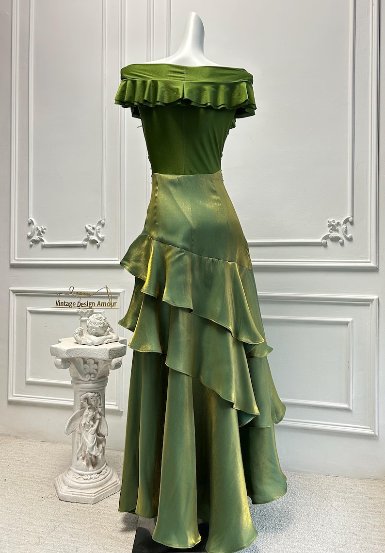 May include: A green off-the-shoulder dress with a ruffled neckline and tiered skirt. The dress is displayed on a mannequin. The text "Vintage Design Amour" is visible on a sign in the background.