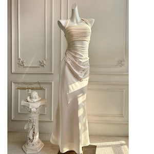 May include: A cream-colored, floor-length gown on a mannequin. The dress features a halter neckline and a ruched bodice with a cutout. The skirt is a flowing, A-line style. A small, decorative pedestal with a sculpture is in the background.