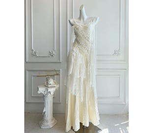 Puede incluir: Un vestido color crema, largo hasta el suelo, sobre un maniquí. El vestido presenta una falda en capas y un corpiño con delicados adornos. Una tela transparente y drapeada cae de los hombros. El vestido se exhibe frente a una pared blanca.