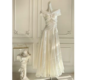 Puede incluir: Vestido color crema con hombros descubiertos y falda fluida. El corpiño presenta un diseño fruncido y delicados detalles de encaje. El vestido se exhibe en un maniquí frente a una pared blanca.