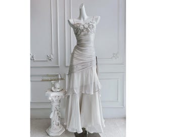 Puede incluir: Vestido blanco largo en un maniquí. El vestido presenta un corpiño fruncido con adornos florales y una falda fluida en capas. Una faja drapeada acentúa la cintura. El fondo es una pared blanca con molduras decorativas.